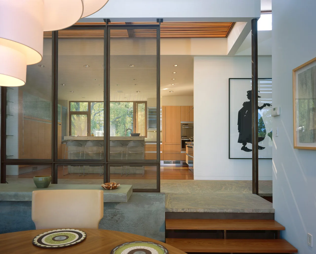 Open kitchen visible through black-framed glass wall from adjacent dining space with wooden table and seating.