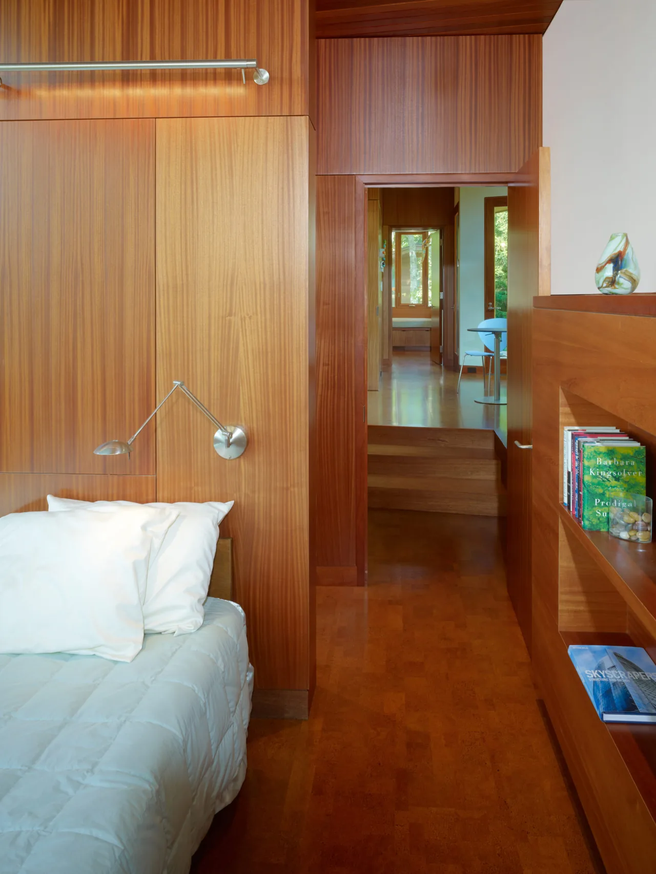 Wood-paneled bedroom interior with built-in shelving and a view through an open door to an adjacent room with natural light.