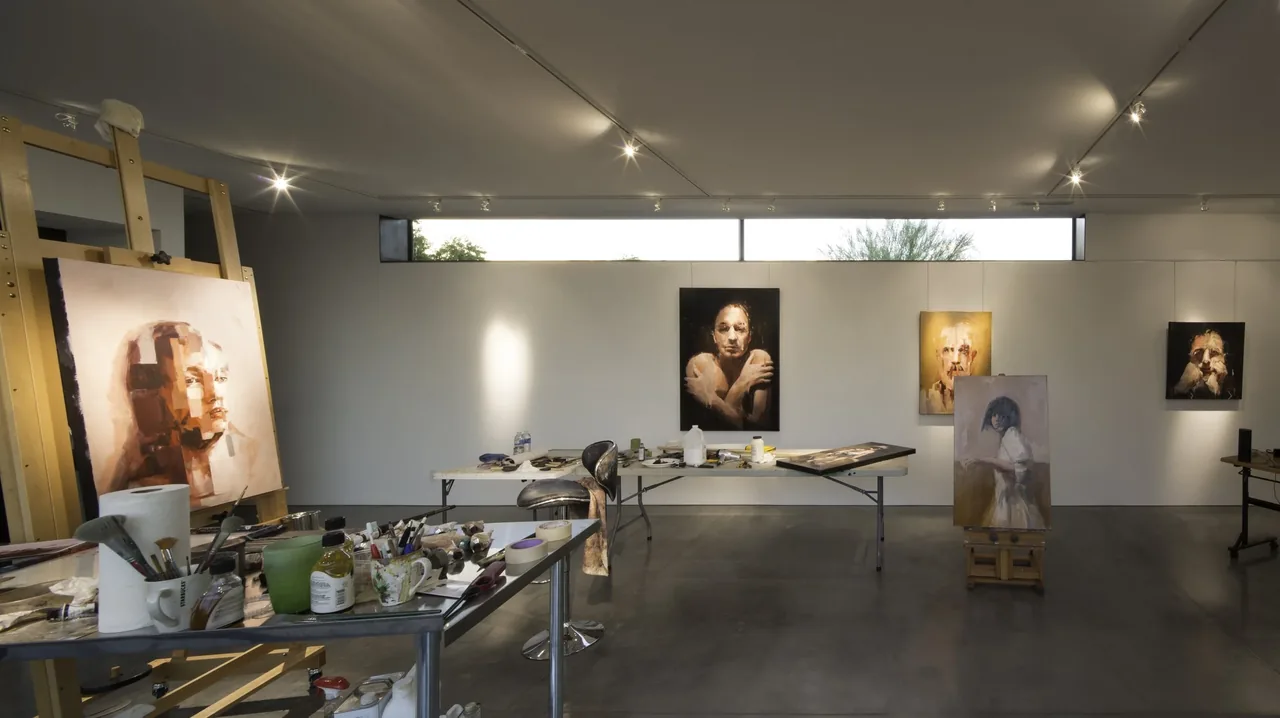 Art studio space featuring portrait paintings on white walls and scattered painting materials on tables.