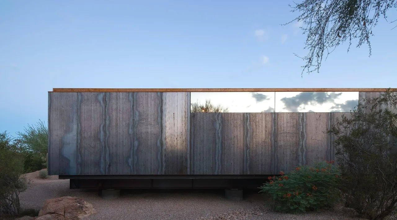 Modern building with a long horizontal mirror-like window and weathered wood panel siding in a desert setting.