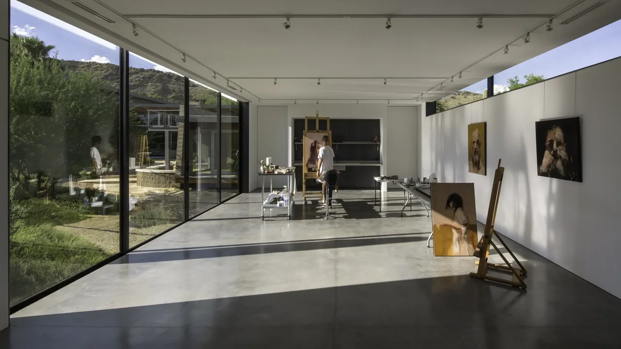 Art studio space with expansive floor-to-ceiling glazing and concrete floor, showing an artist working inside.