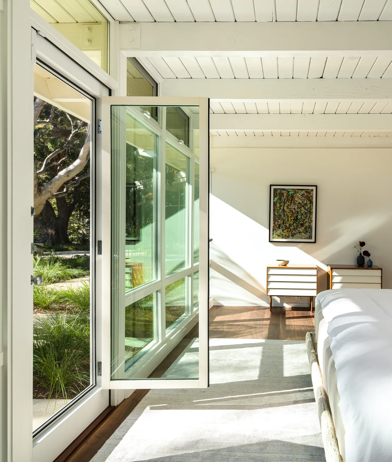 Bedroom with white beamed ceiling, wood floor, white-framed glass door opens to landscaped garden outside.