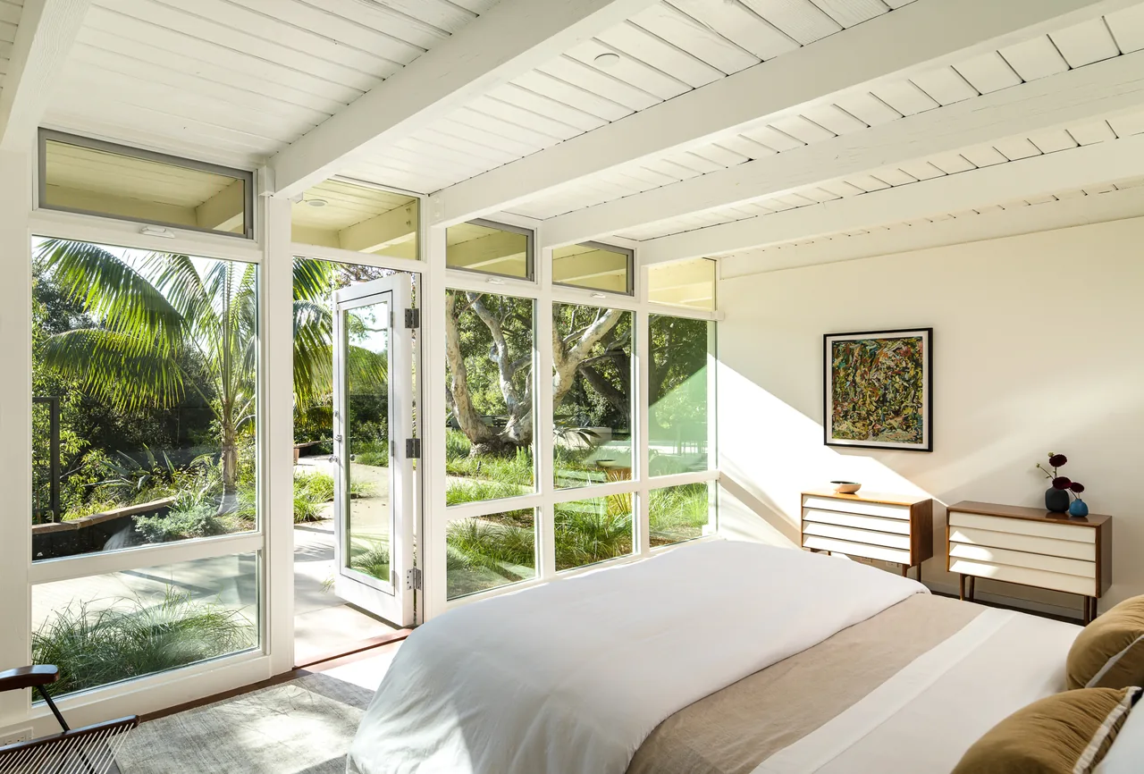 Veneklasen House bedroom features floor-to-ceiling white framed windows and a glass door leading to a landscaped garden.