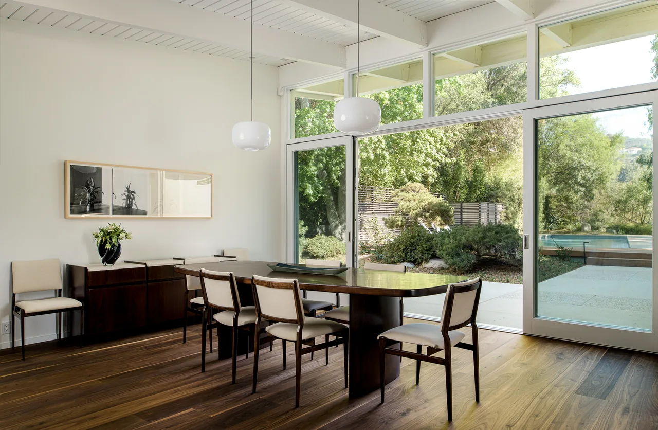 Veneklasen House dining room features a wooden table with upholstered chairs and large windows overlooking a garden and pool.