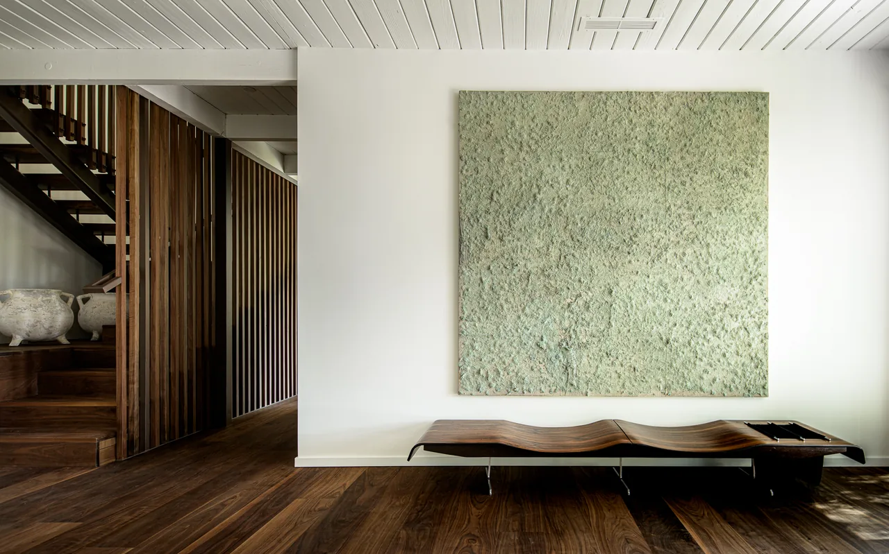 Minimalist interior with dark wood flooring, a sculptural wave bench, and textured abstract wall art under a white beamed ceiling.