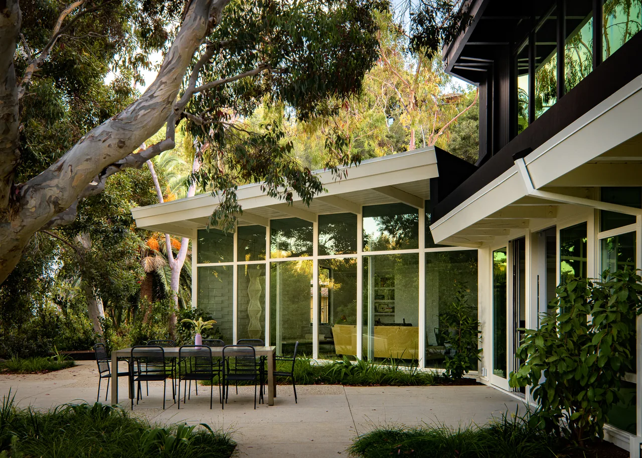 Two-story modern house with floor-to-ceiling glazing, a sloped roof, and an outdoor dining area in a landscaped setting.