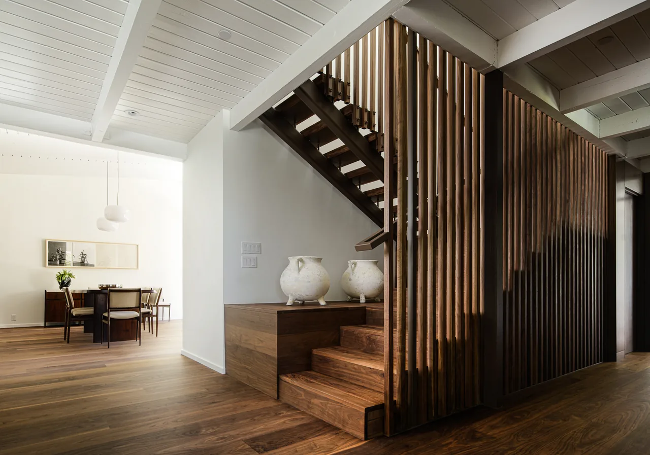 Interior view showing a wooden staircase with vertical slats and a dining area with wooden furniture under a white beamed ceiling.