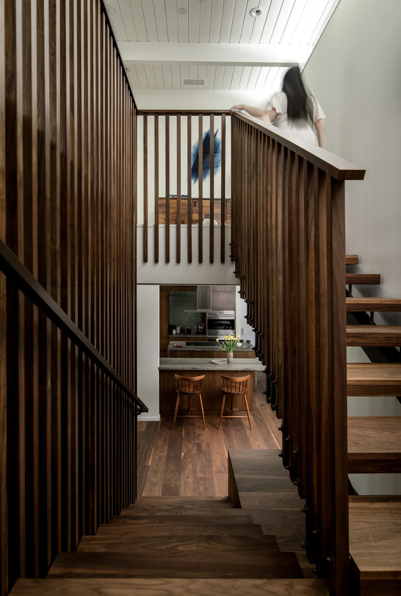 Wood-paneled staircase with vertical railings overlooks the kitchen with walnut floors and wood cabinetry at the Veneklasen House.