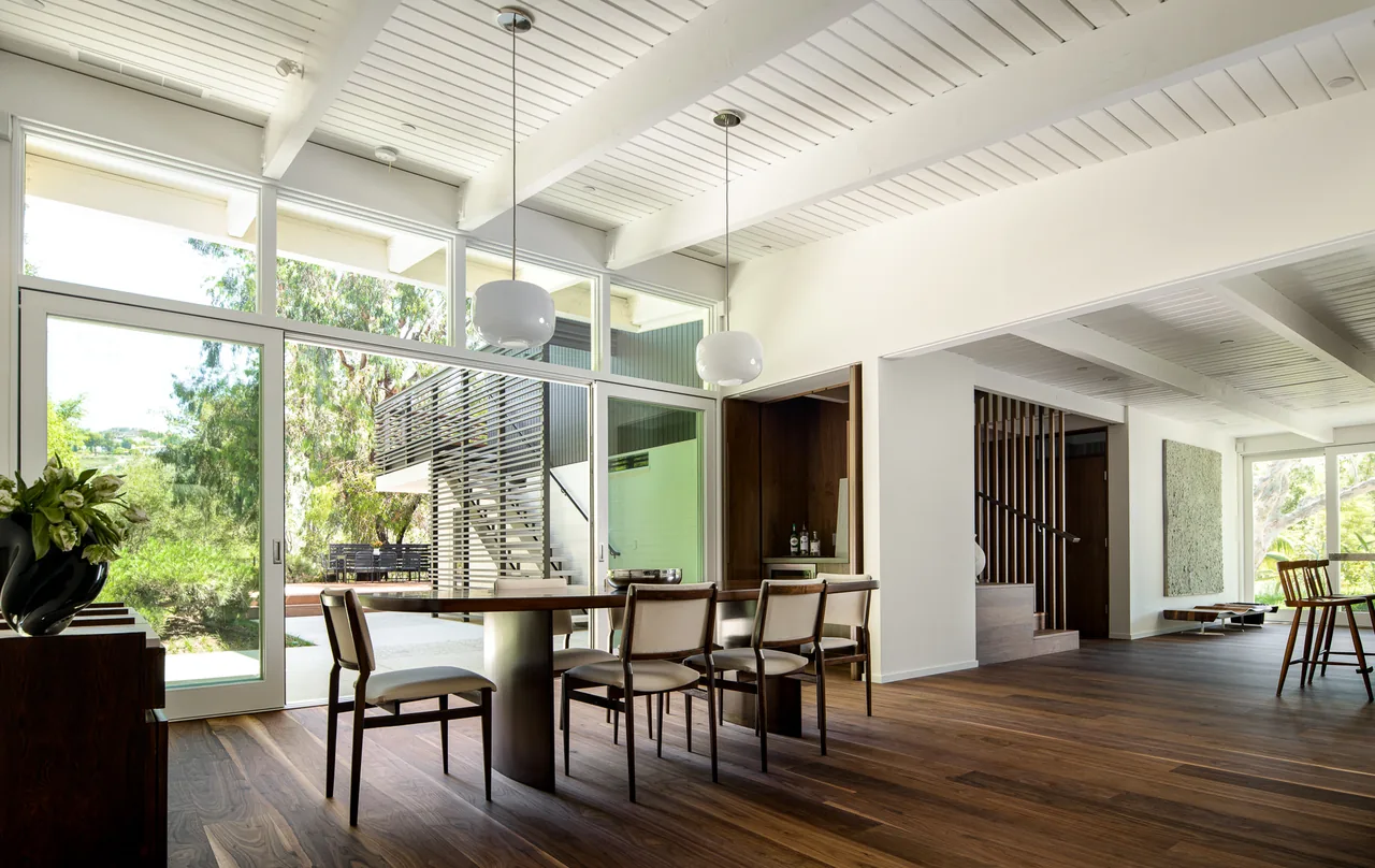 Interior dining space with wood flooring, white beamed ceiling, large sliding glass doors, and modern wooden chairs.