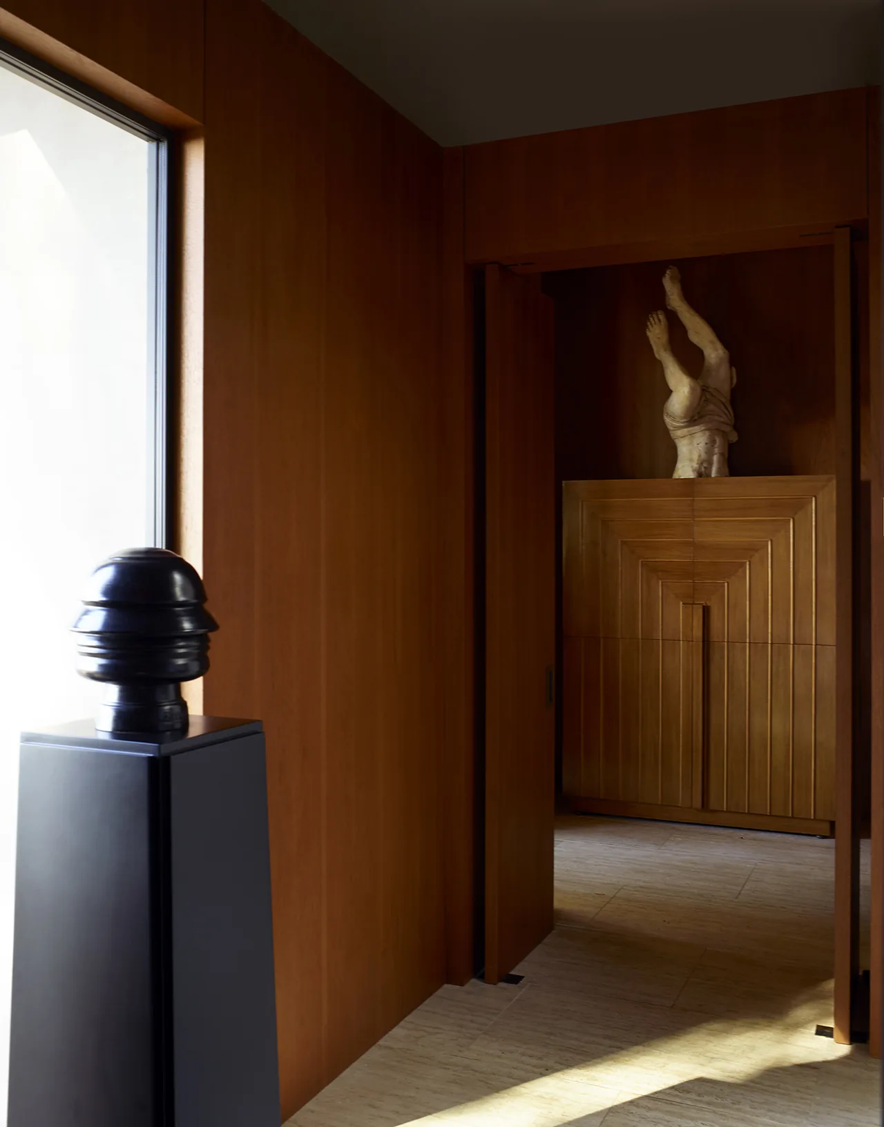 Wood-paneled hallway featuring a sculptural statue atop a carved wooden cabinet with travertine flooring.