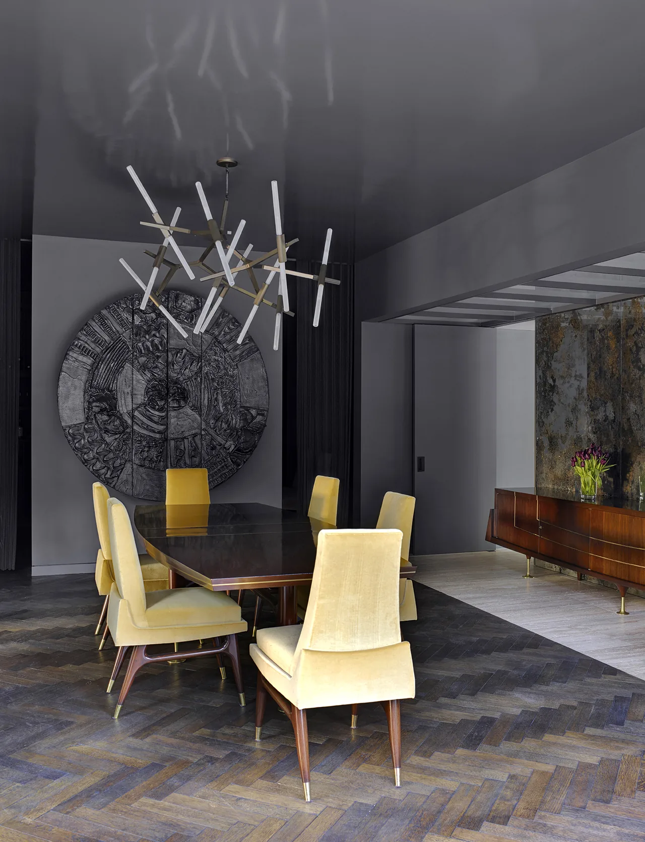 Dining area features a dark wood table, yellow upholstered chairs, and a sculptural chandelier with a large circular artwork on the wall.