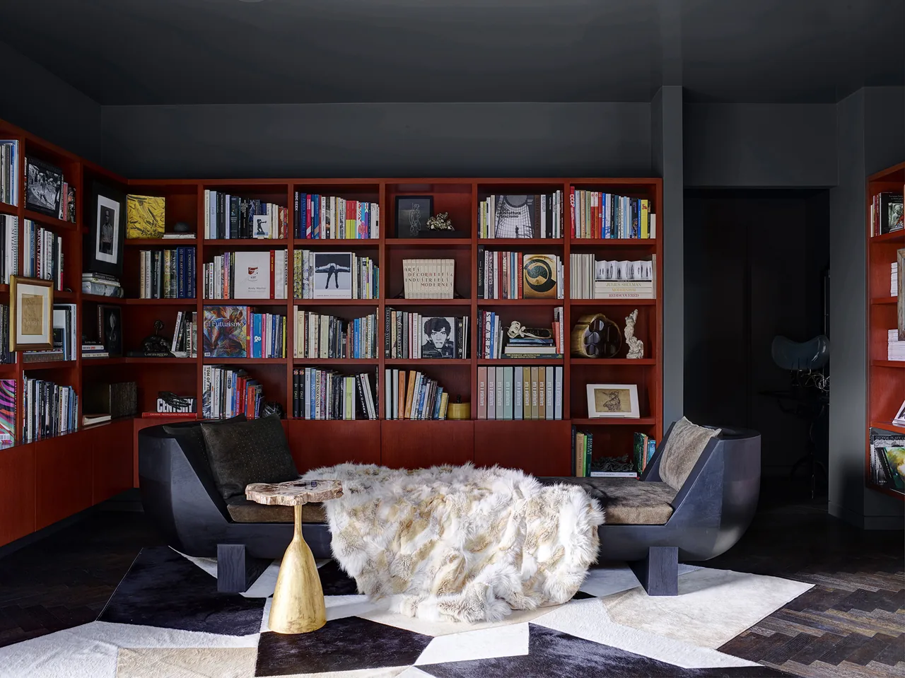 A reading room features dark sculptural seating, contrasting geometric rugs, and built-in wood bookshelves filled with books and decor.