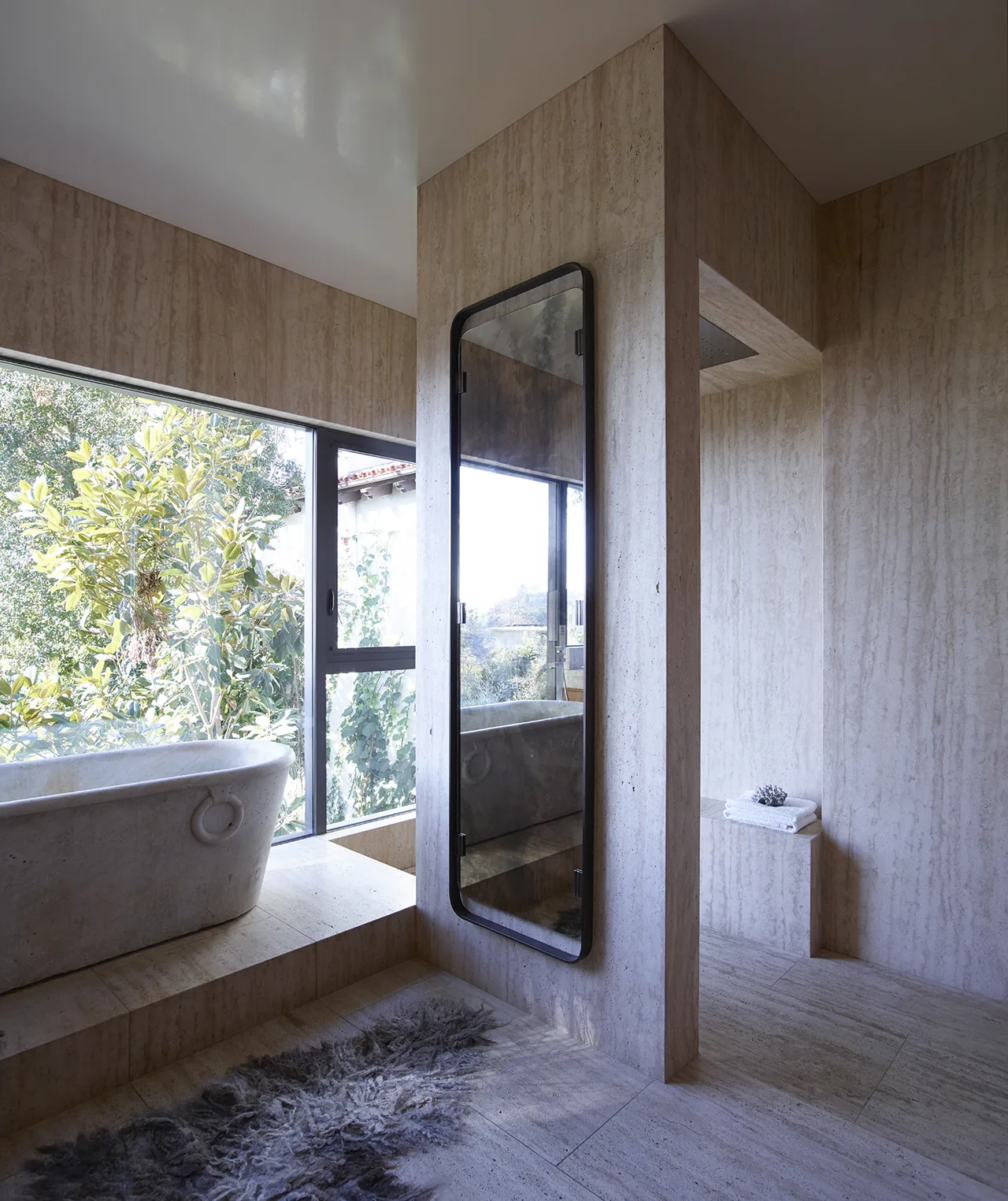This bathroom features travertine surfaces, a freestanding tub by a window, and a vertical mirror with a black frame.