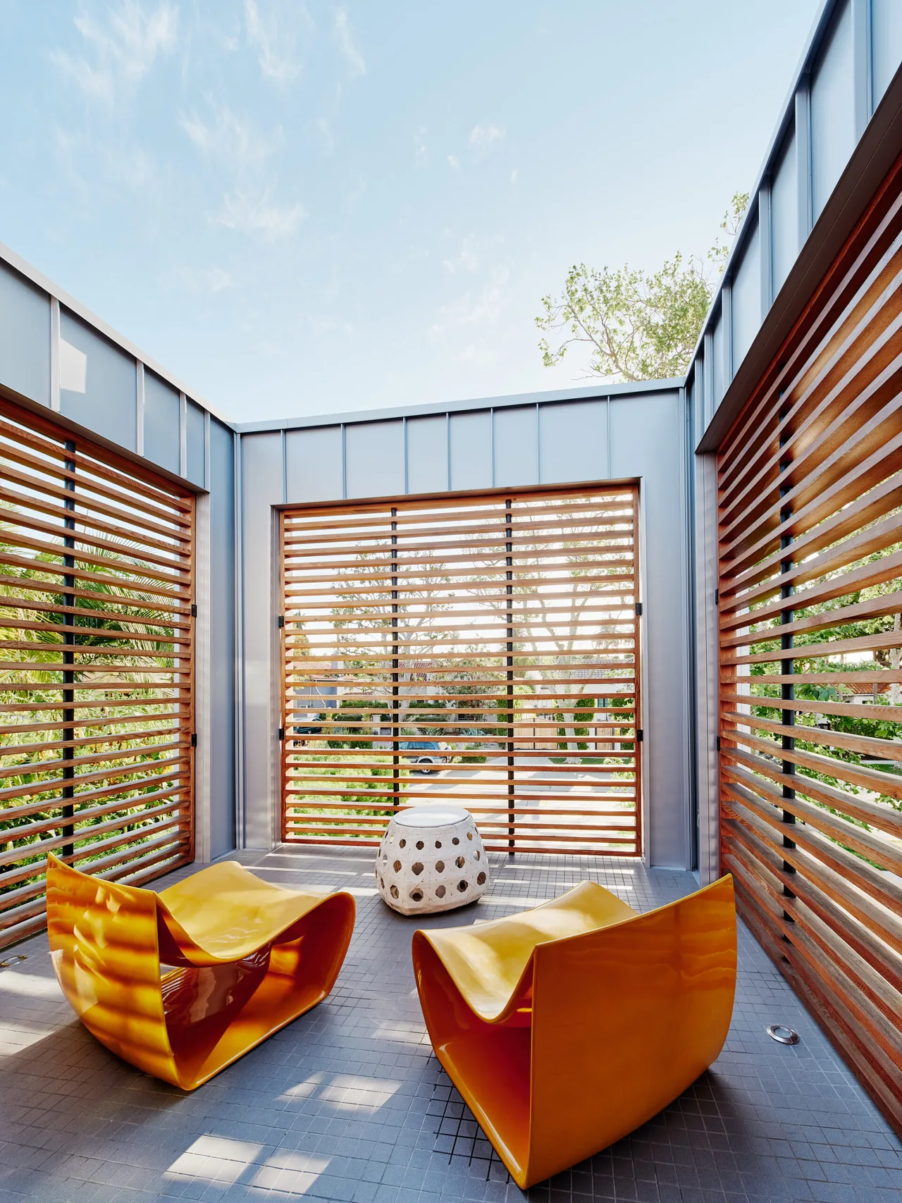 A courtyard space features yellow sculptural chairs and a small white side table enclosed by wood slat walls and an aluminum frame.