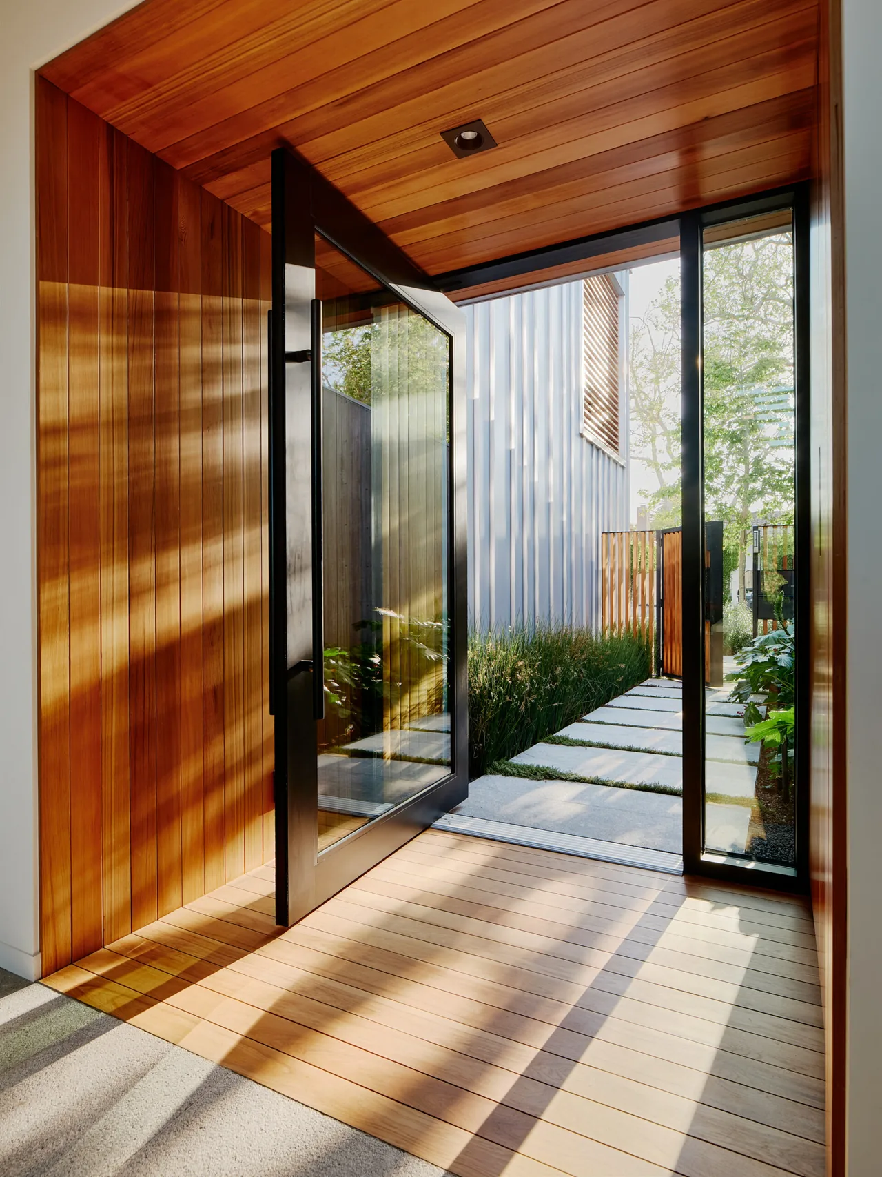 Entryway features a pivot door with black frames and wood panel cladding indoor and out in this Los Angeles residence.