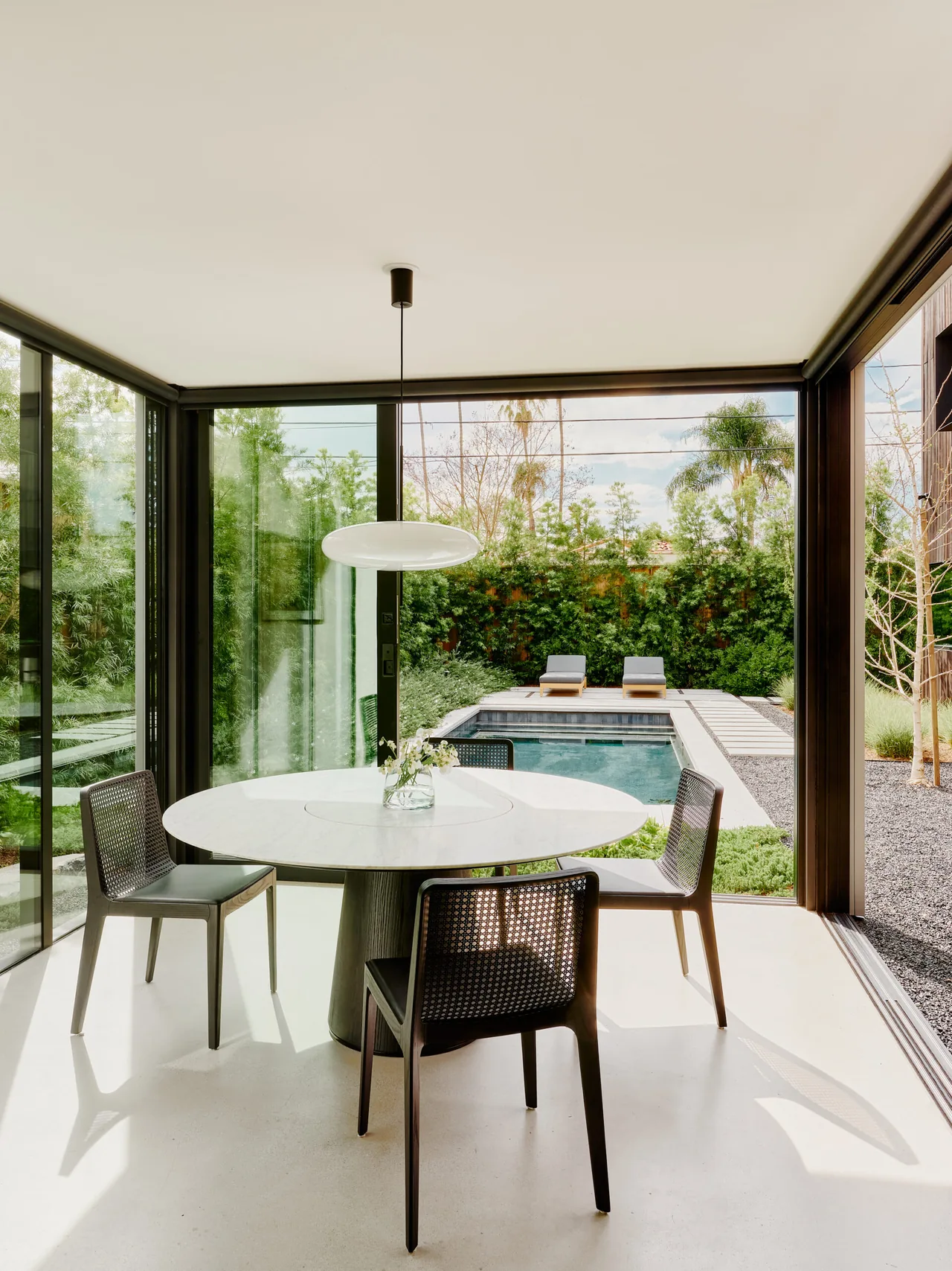 Dining space with round table and black chairs opening to a garden and pool via large glass walls.