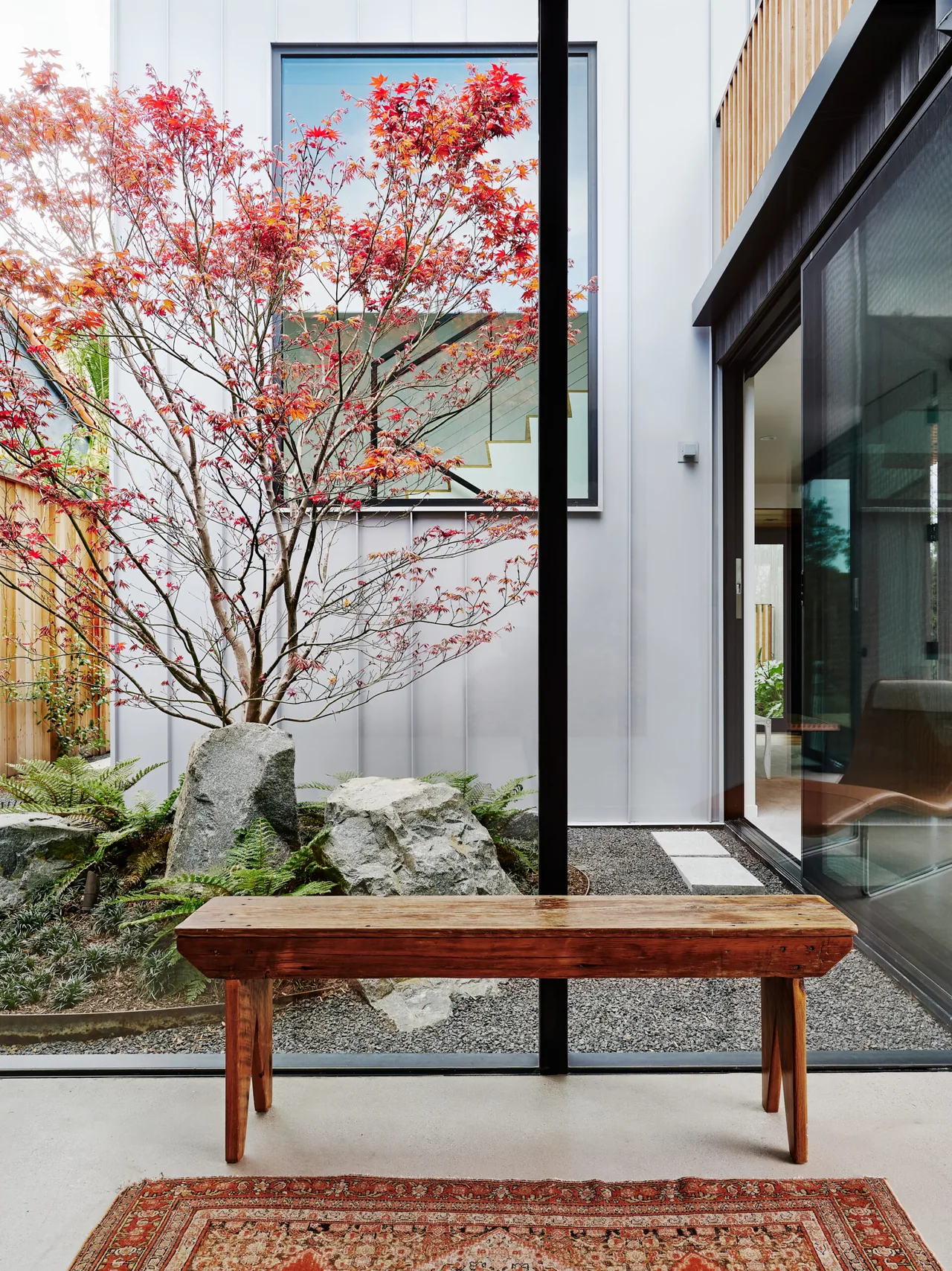 View from indoors showing a wooden bench facing a landscaped courtyard with a red-leafed tree and large rocks.