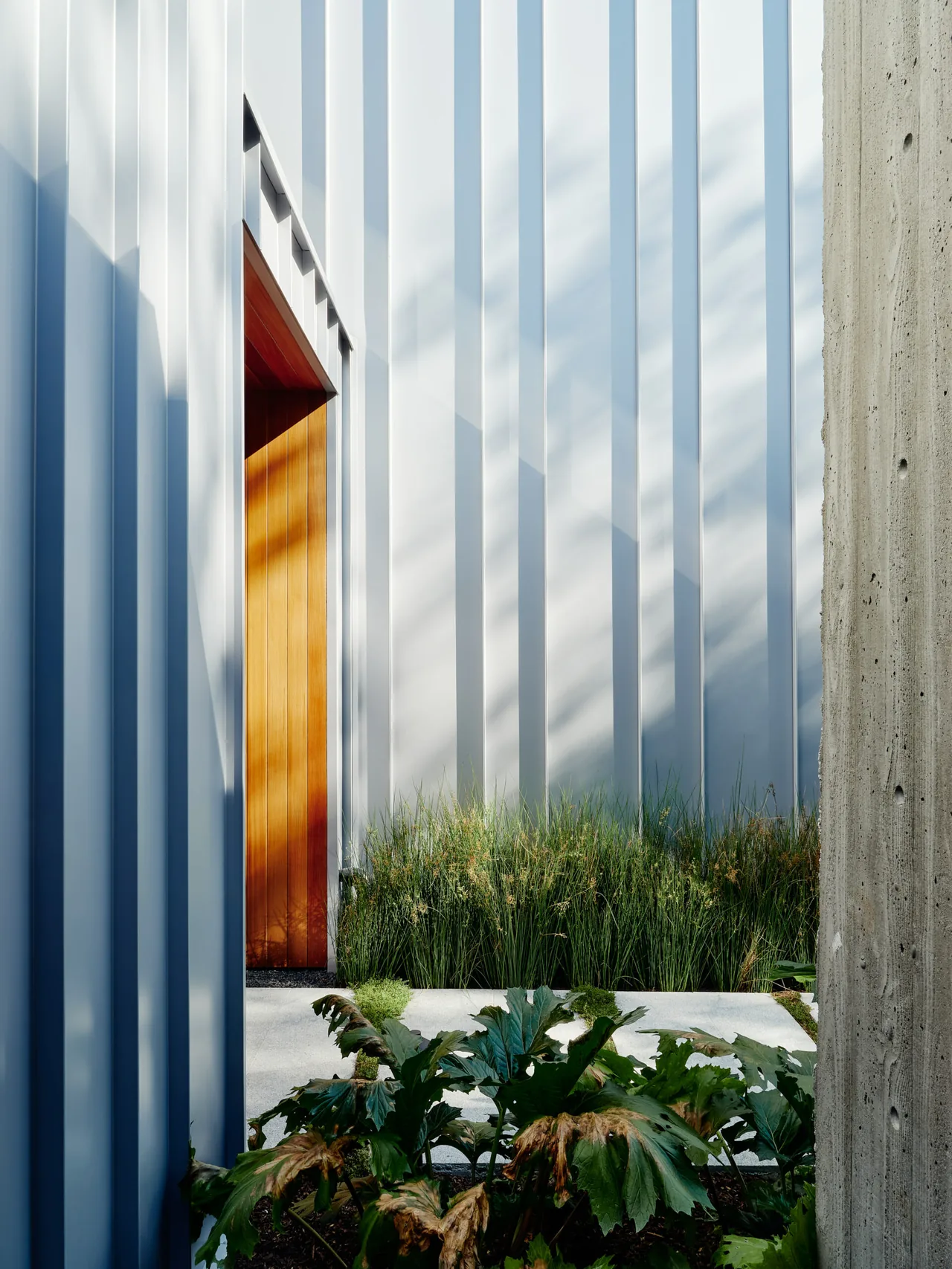 A corrugated metal exterior wall with a wooden door and nearby concrete pillar in the Los Angeles residence by Clive Wilkinson Architects.