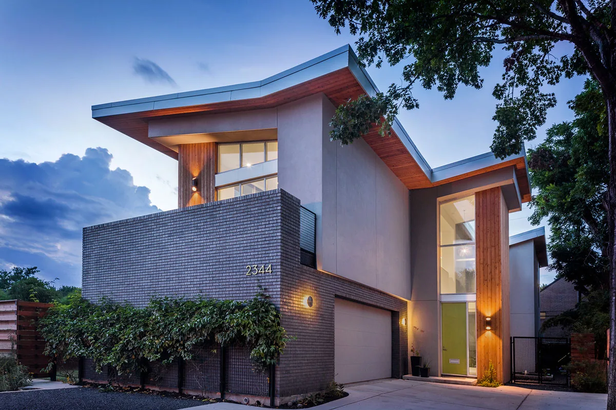 HOUSE 2344 features a contemporary exterior with angled metal roofs, wood siding, and large windows at dusk in Houston.