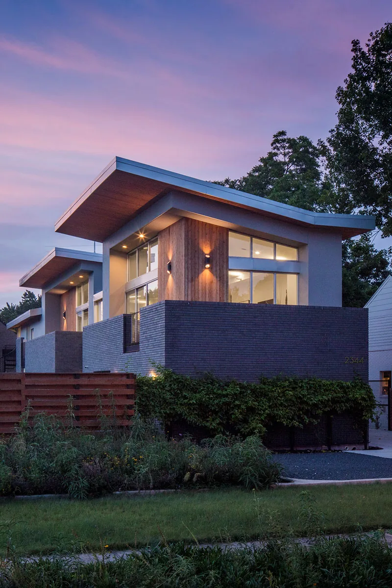 HOUSE 2344 features angled overhanging rooflines with wood and brick siding, illuminated by warm exterior lights at dusk.