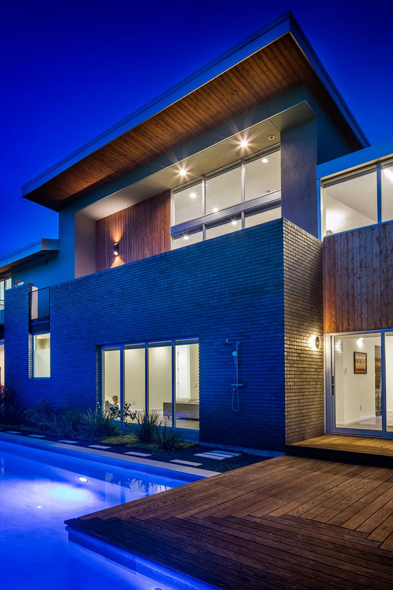 Two-story modern house with brick and wood exterior and a pool illuminated in blue light at dusk.
