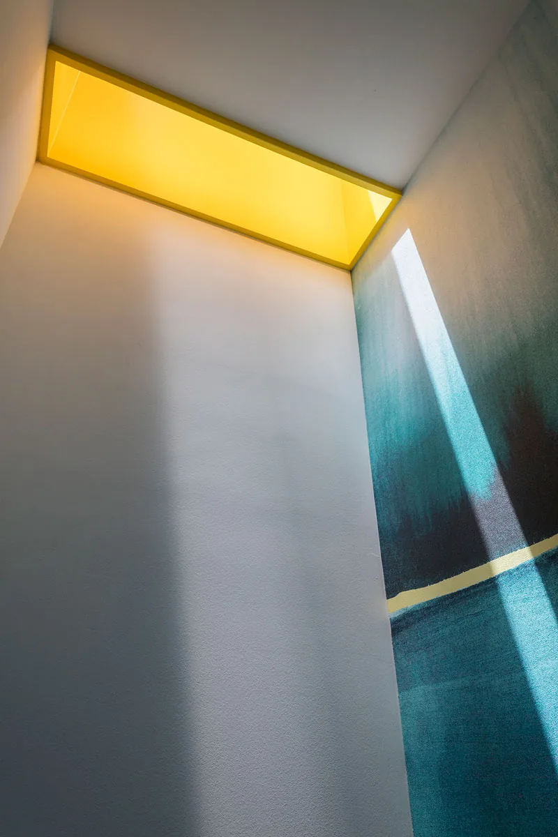 A yellow-framed skylight brings natural light into a narrow interior corner with a blue accent wall.