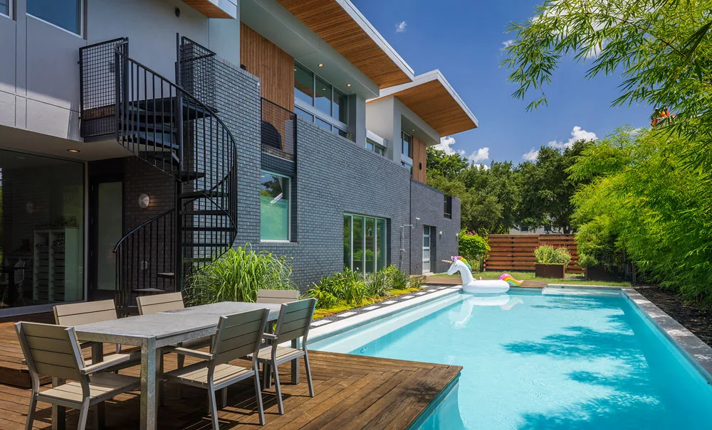Modern two-story home with a gray brick exterior, wooden deck dining area, spiral stair access, and a pool with a unicorn float.