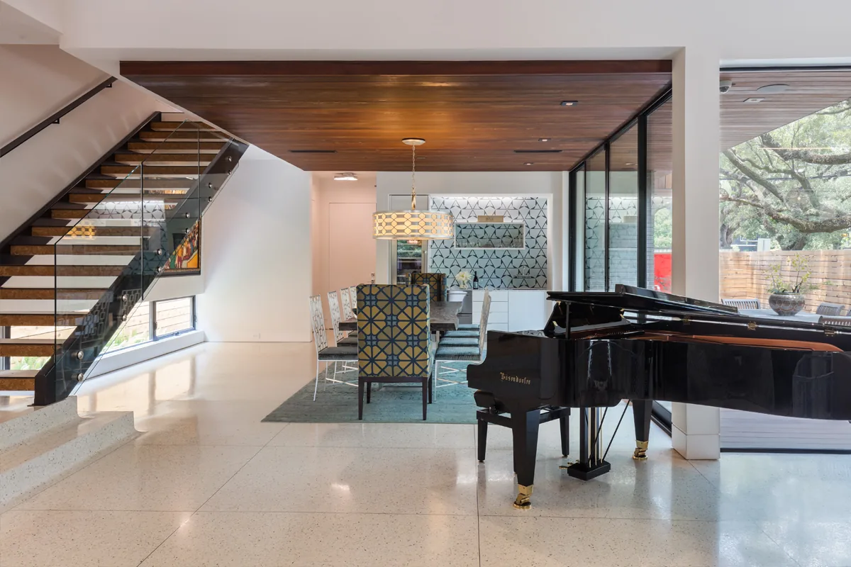 Interior view showing the dining area, staircase with glass railing, and black grand piano next to floor-to-ceiling windows.