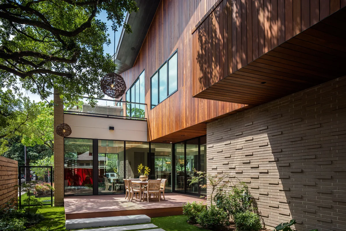 The exterior features a wooden-paneled upper facade, a textured brick wall, and a patio dining area connected to the interior by glass walls.