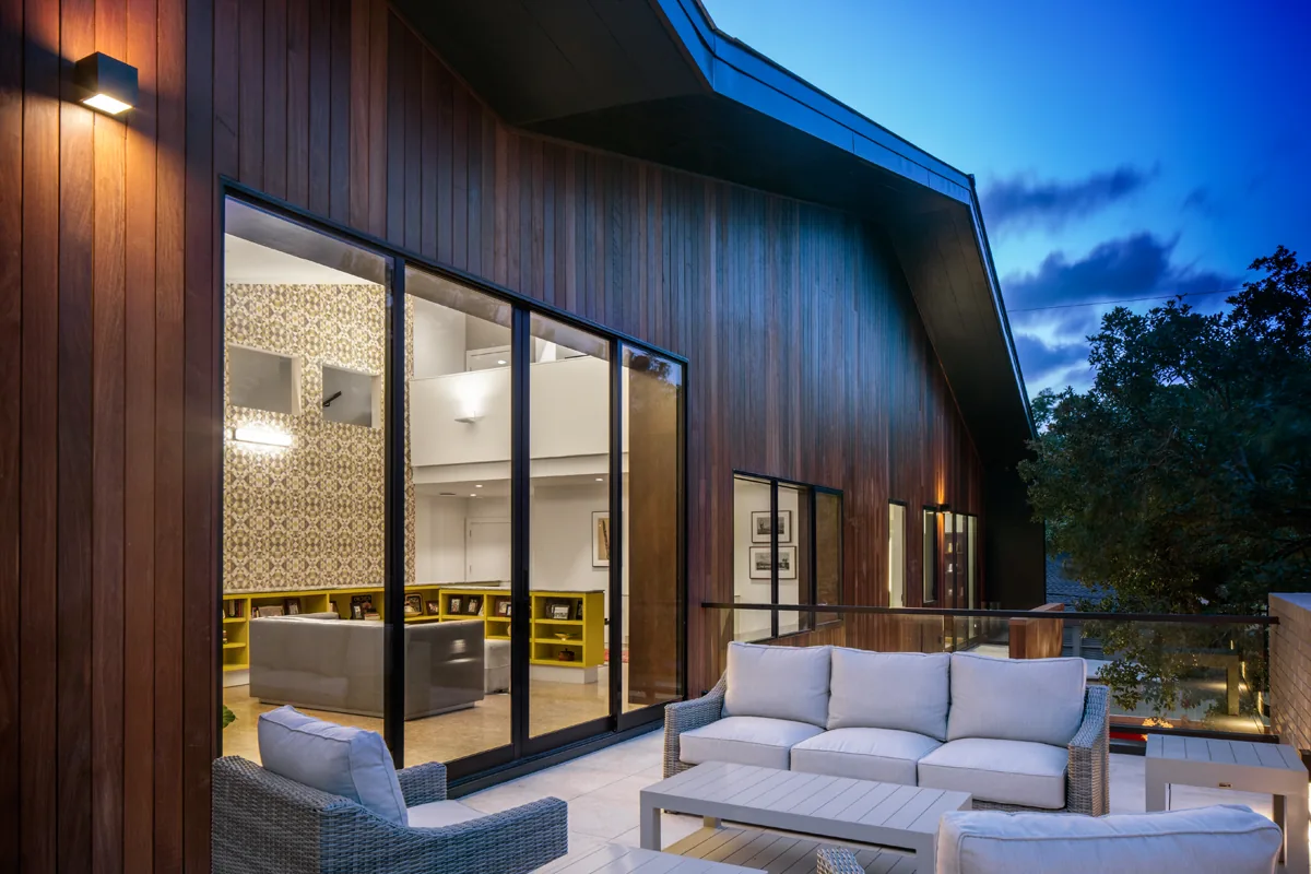 Balcony with cushioned wicker seating outside a wood-paneled home with large glass sliding doors connecting to the interior living space.