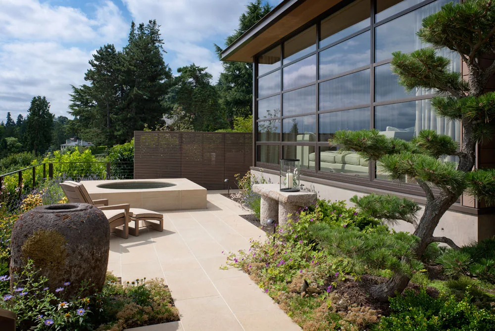 Patio area features a built-in spa, stone tile floor, and large multi-pane windows reflecting greenery.