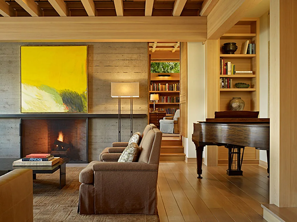 Living area featuring concrete fireplace, wooden beams, brown armchairs, and a grand piano with built-in wooden shelving.