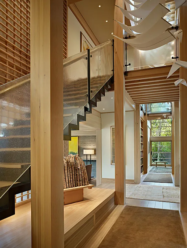 Interior view of a wood post-and-beam structure with a floating staircase and hall leading to a light-filled room.