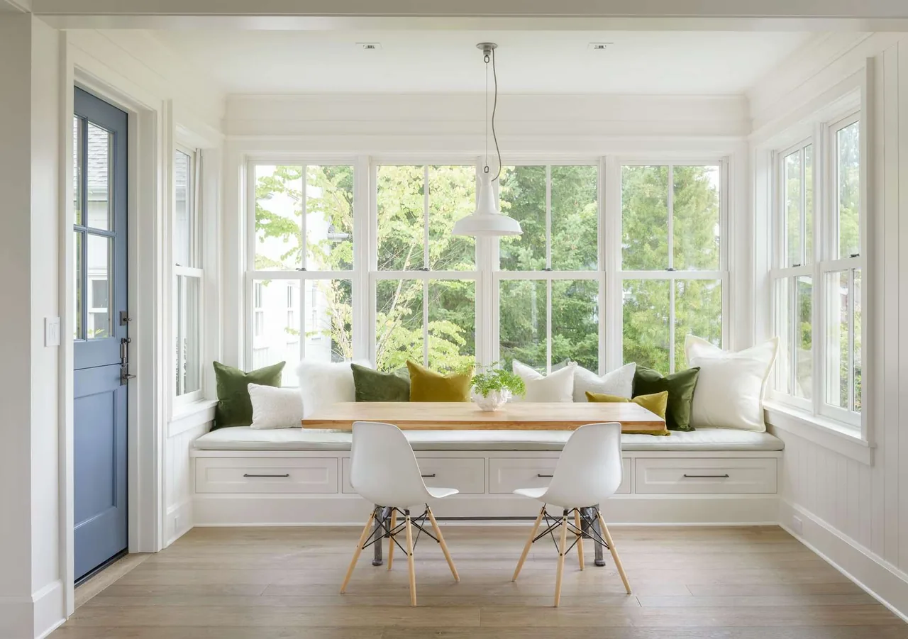 Dining nook with built-in bench, wooden table, and white chairs under pendant light in a bright room with many windows.