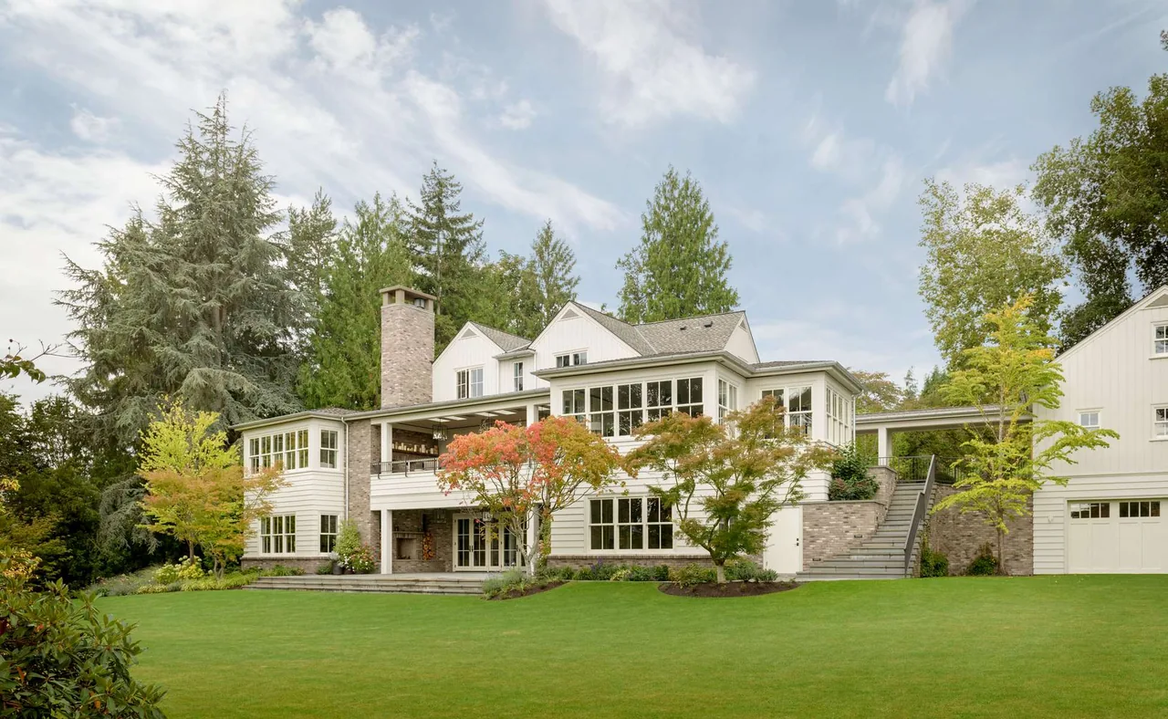 Rear view of a modern farmhouse with white wood siding, numerous large windows, a stone chimney, and landscaped lawn in Seattle.