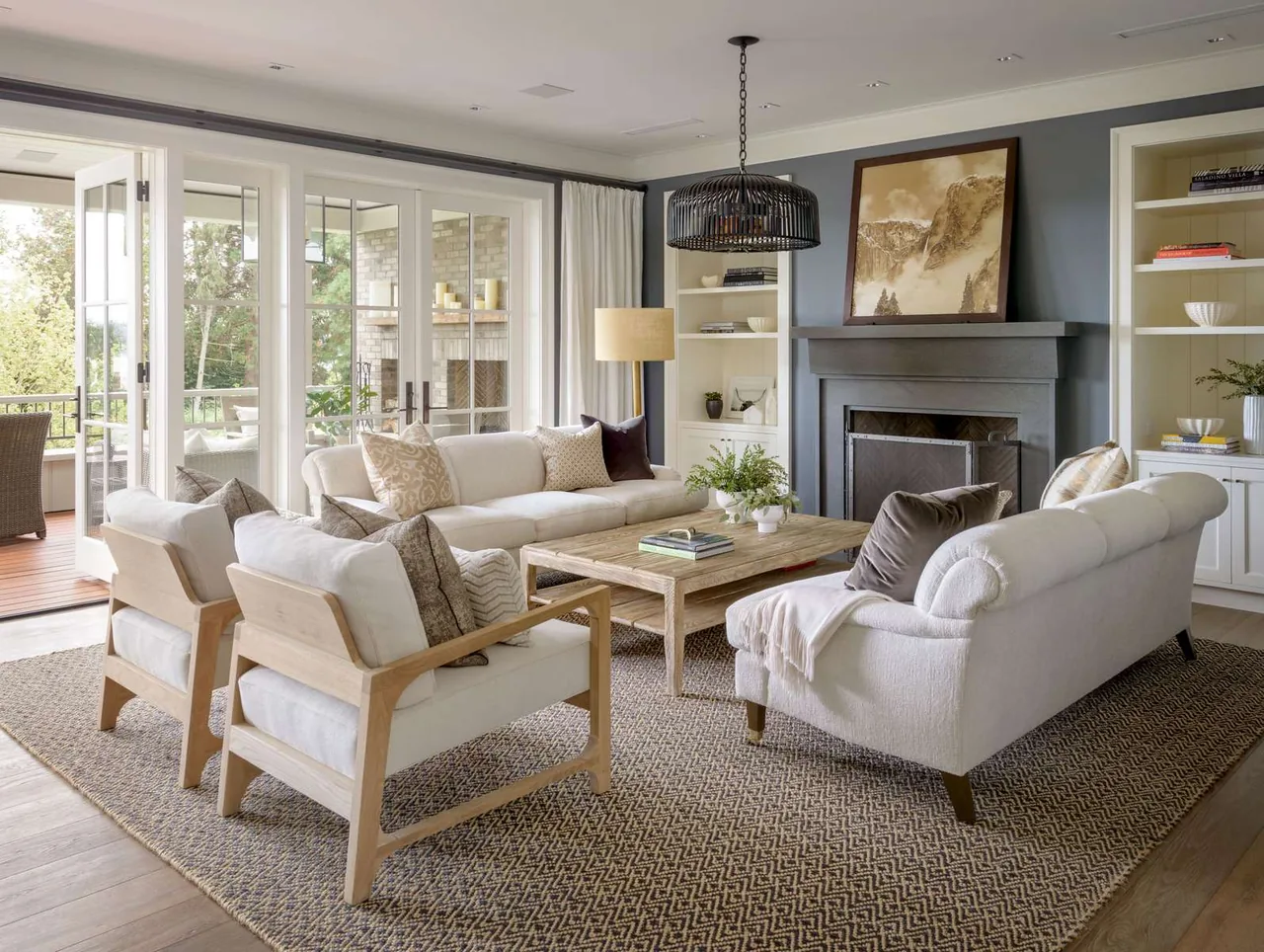 Modern farmhouse living room features neutral sofas, wooden armchairs, a patterned rug, and a gray fireplace with built-in shelving.