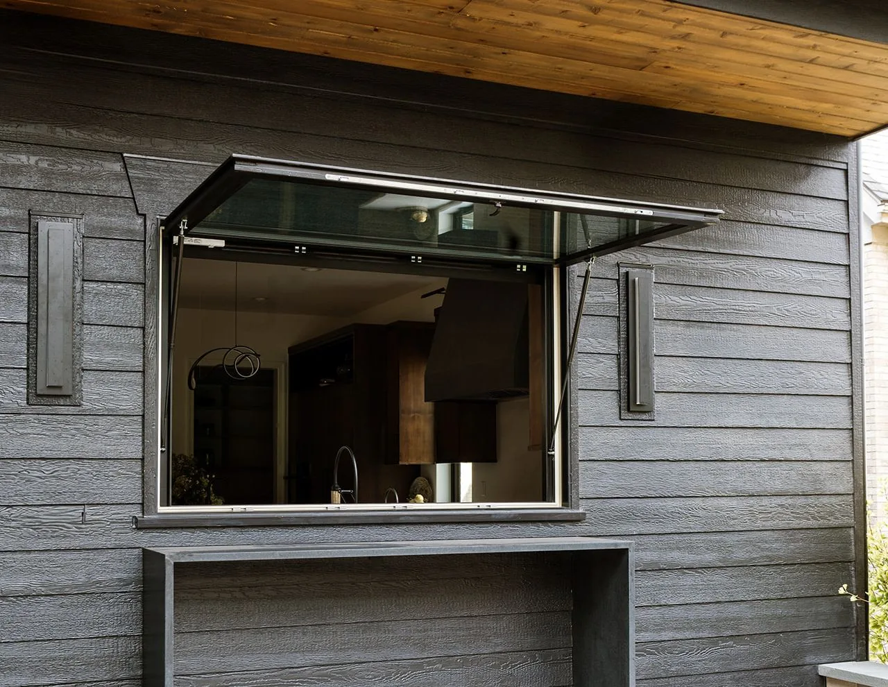 This Clarendon Hills Residence by Charles Vincent George Architects features a black-framed awning window set in black wood siding with a wooden ceiling overhang.