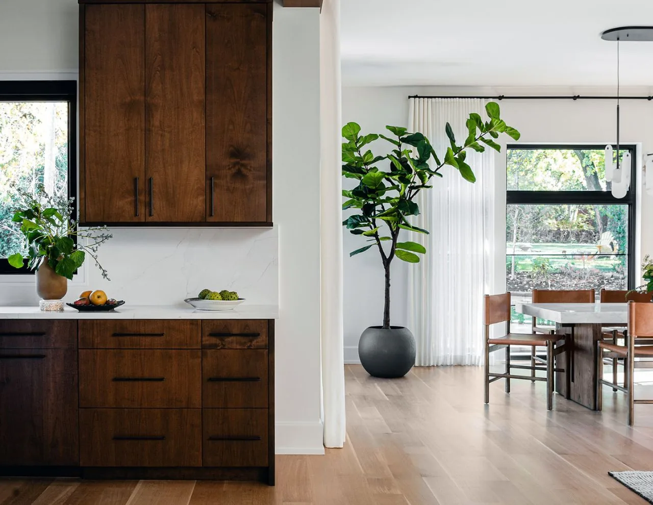 Kitchen features dark wood cabinets with a marble backsplash adjacent to a dining space with a large potted plant and natural light from black-framed windows.