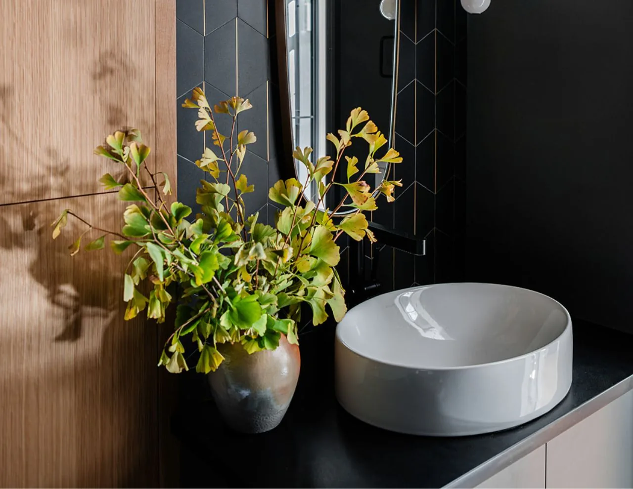 A modern bathroom counter features a white round vessel sink and a vase with green foliage on a black countertop.