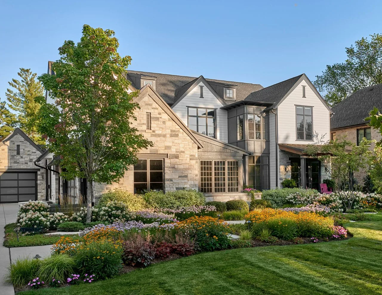 This transitional farmhouse residence features multiple gables, dormer windows, and a lush landscaped garden seen from the front exterior.