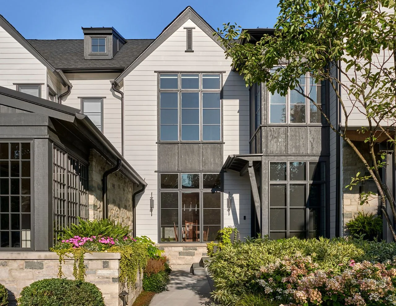 The transitional farmhouse residence features white horizontal siding, large black-framed windows, and stone detailing on the exterior.