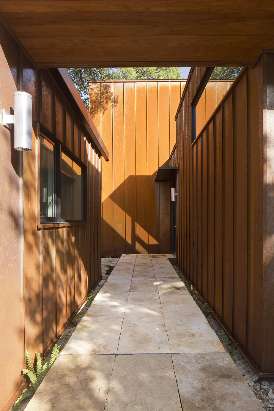 Outdoor passageway between buildings clad in rust-colored metal panels with stone paving and wall-mounted lights.