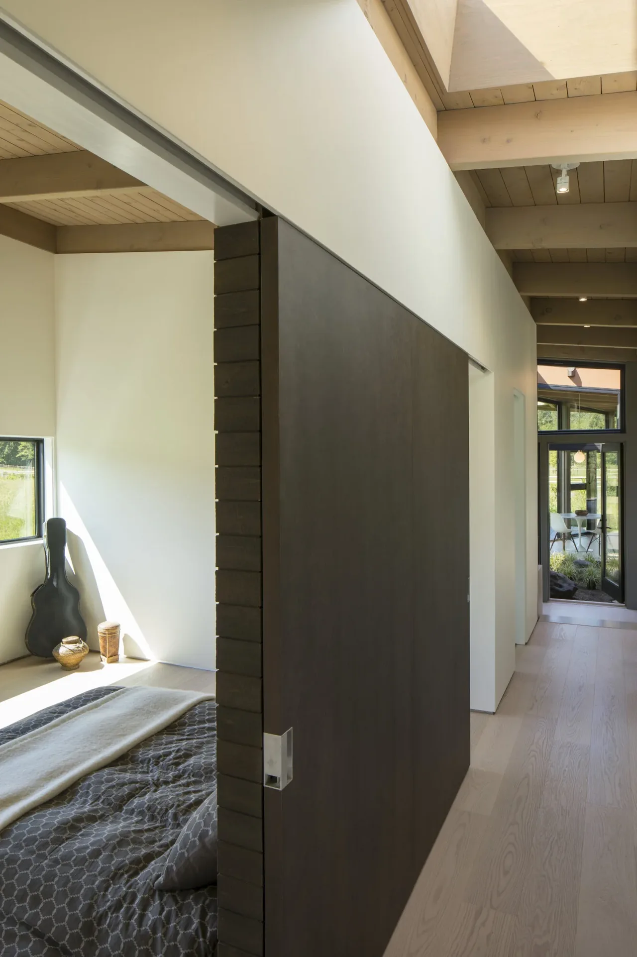 Sliding dark wood door separates the hallway from the bedroom that has wood beam ceiling and light wood flooring.