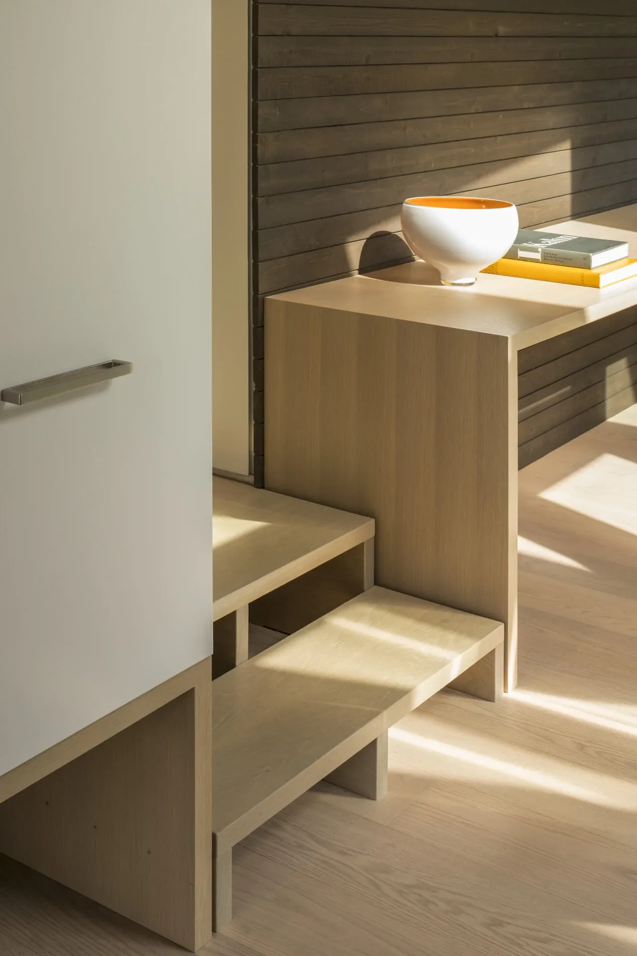 Minimalist indoor seating area with built-in light wood bench steps against dark horizontal wood panel wall.