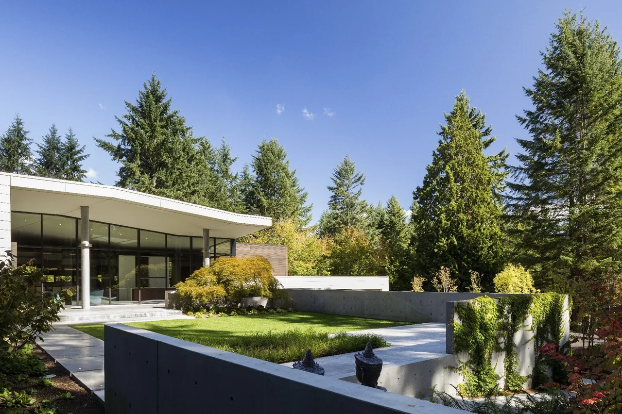 The Inside Outside House features large glass windows and a flat roof surrounded by landscaped greenery and concrete retaining walls.