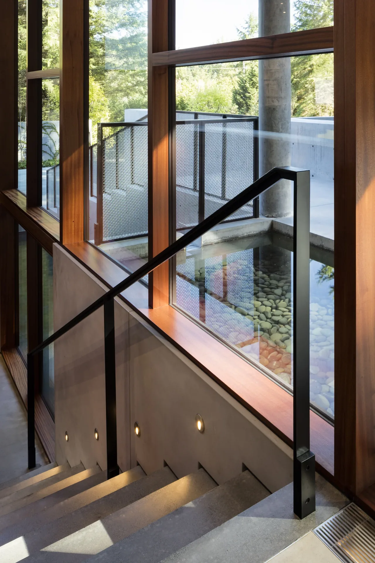 Stairwell with concrete steps and black metal handrail next to floor-to-ceiling wooden framed windows overlooking a pebble-filled water feature and patio.