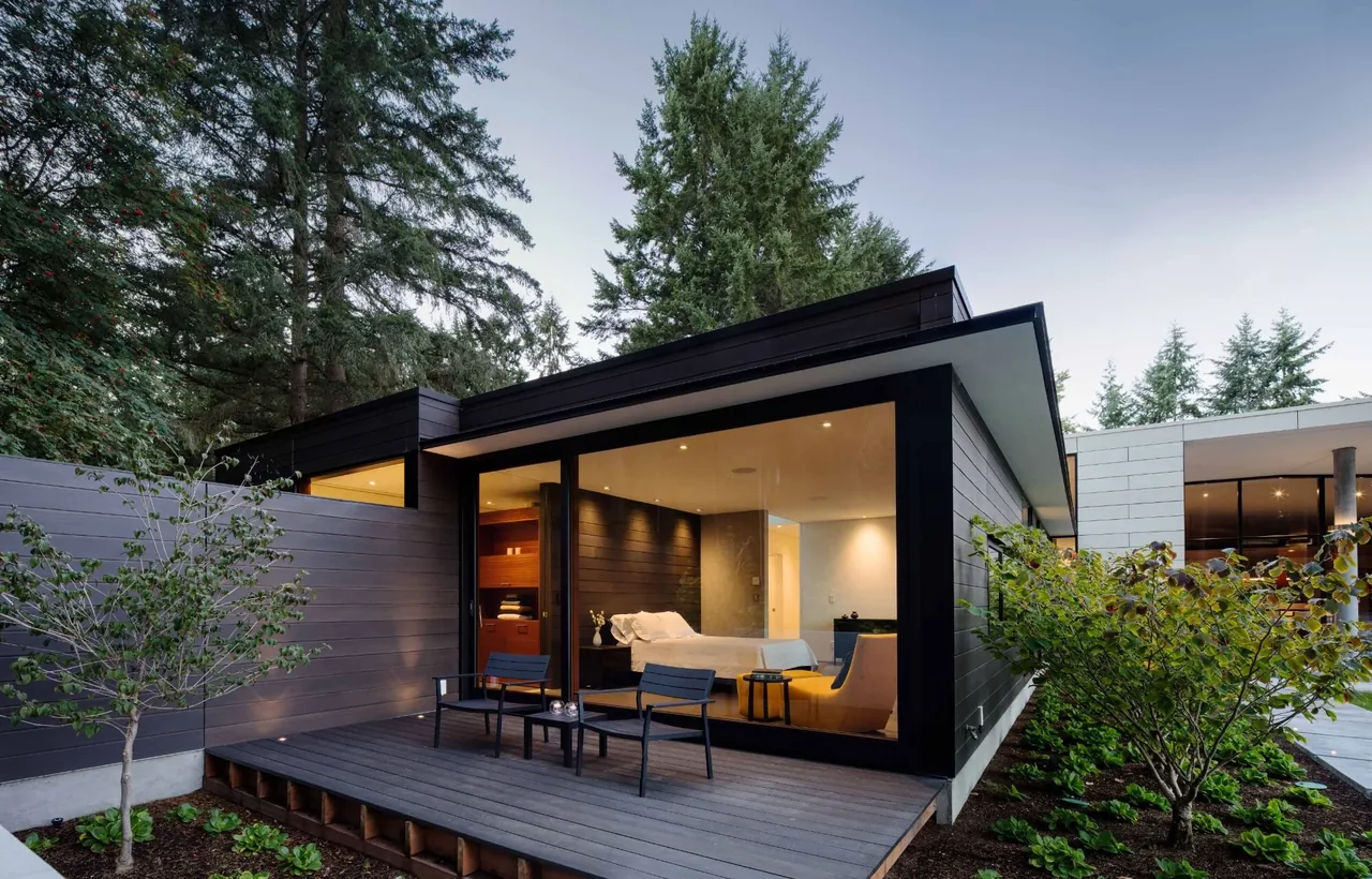 The Inside Outside House by David Coleman Architecture features dark wood siding, large corner windows, and an outdoor deck with seating.