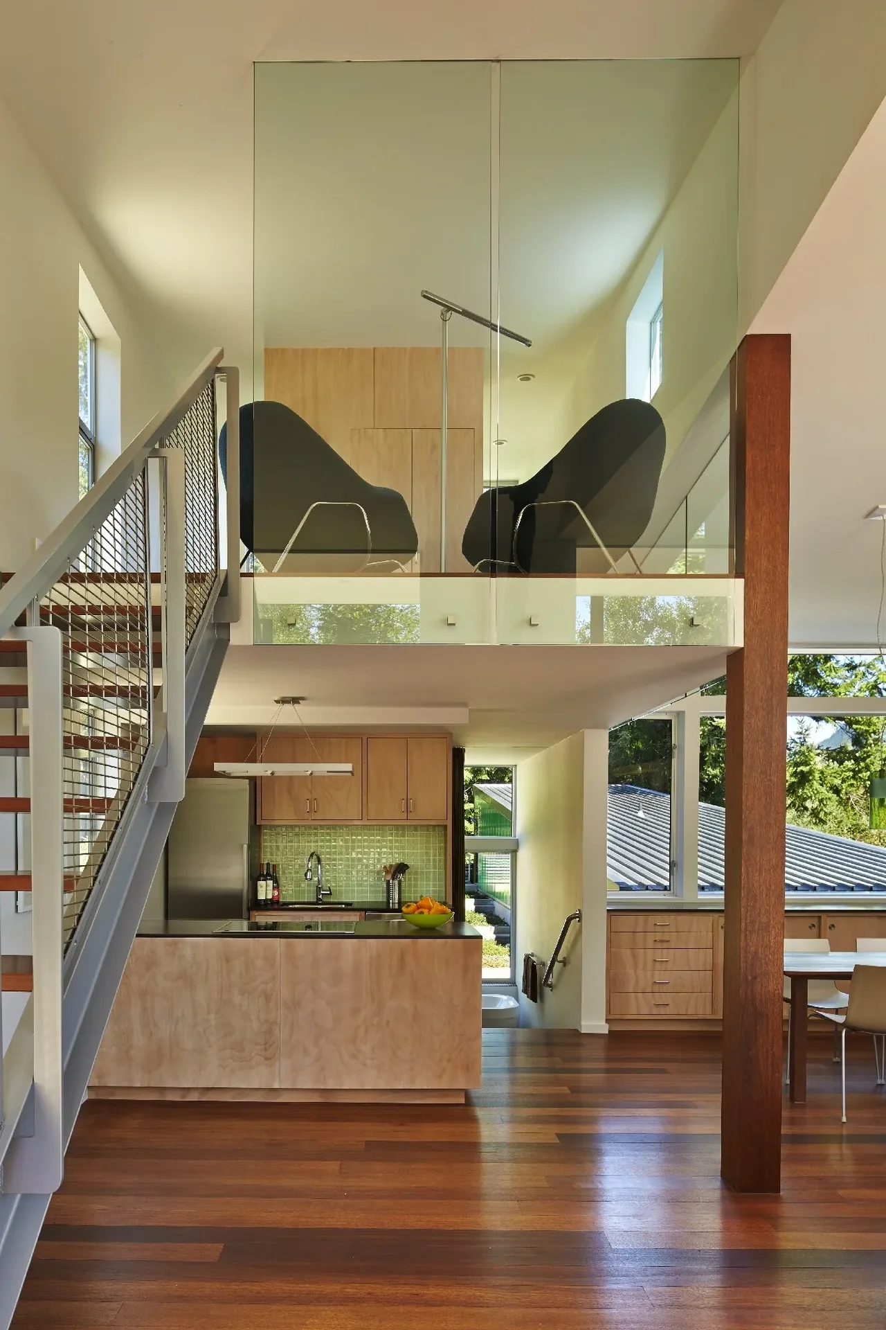 Modern interior showing the kitchen with a wooden island, metal mesh railing staircase, and an upper-level seating area enclosed by glass walls.