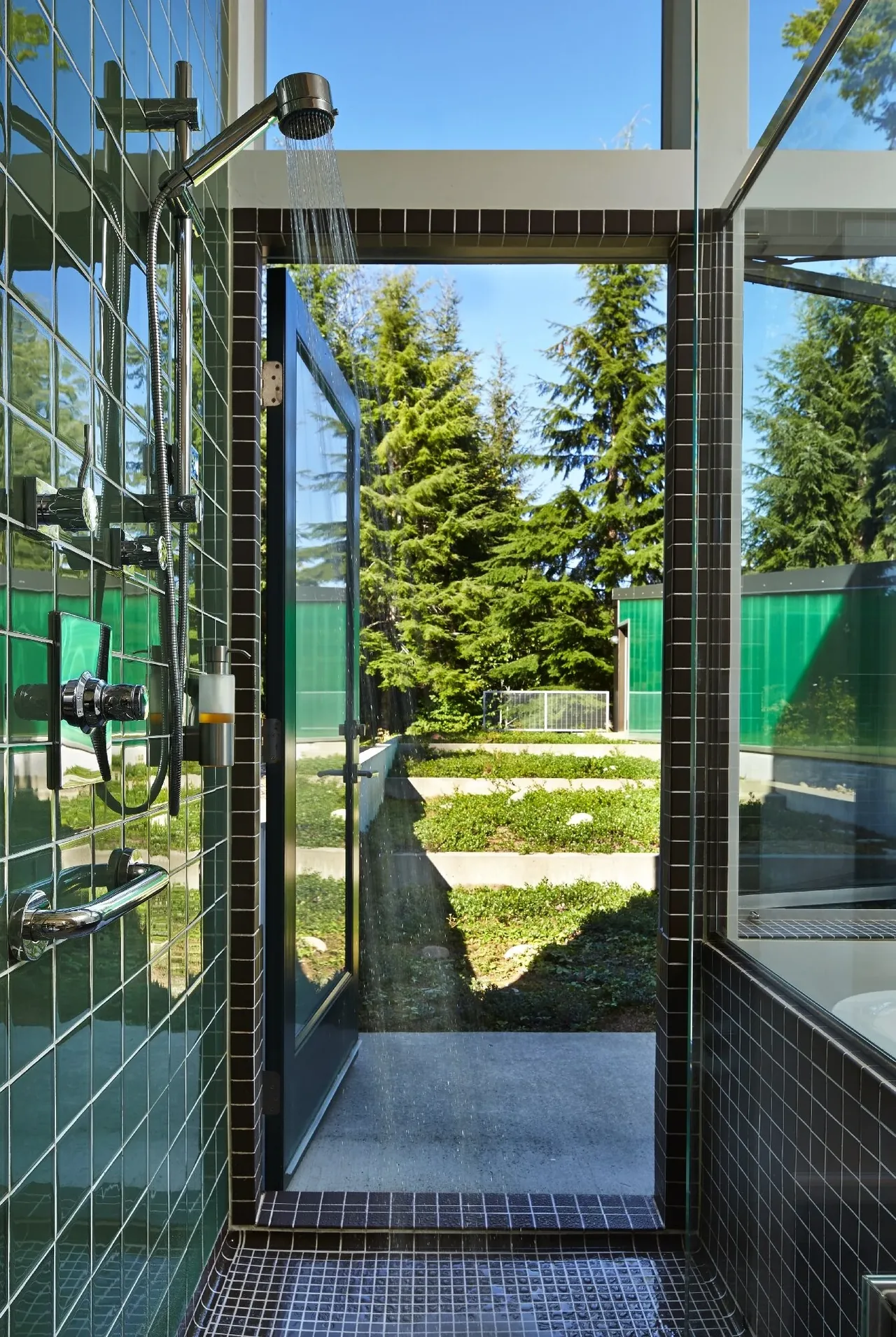Shower with green tile walls opens to a terraced garden outside with forest views.
