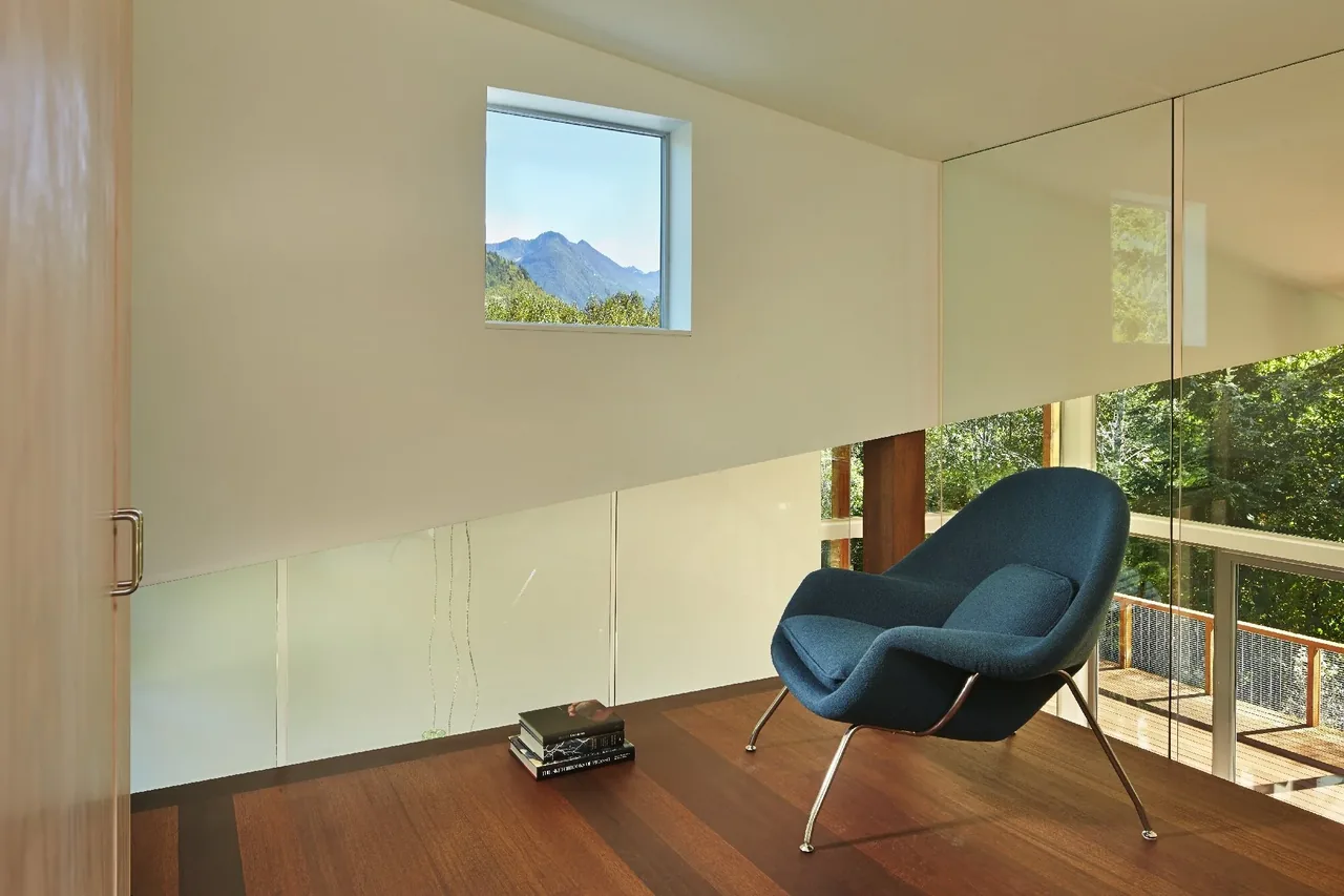 Loft seating area with a blue lounge chair and views of forest and mountains through glass railings and window at Bear Run Cabin.
